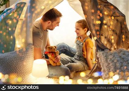 family, hygge and people concept - happy father with teddy bear toy and little daughter playing in kids tent at night at home. happy family playing with toy in kids tent at home