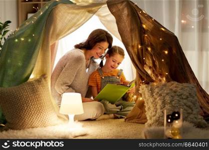 family, hygge and people concept - happy mother and little daughter reading book in kids tent at night at home. happy family reading book in kids tent at home