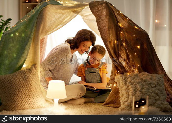 family, hygge and people concept - happy mother and little daughter reading book in kids tent at night at home. happy family reading book in kids tent at home