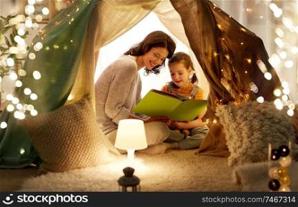 family, hygge and people concept - happy mother and little daughter reading book in kids tent at night at home. happy family reading book in kids tent at home