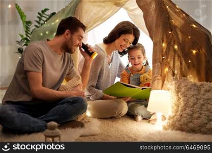 family, hygge and people concept - happy mother, father and little daughter reading book with torch light in kids tent at night at home. happy family reading book in kids tent at home
