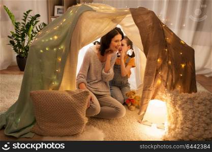 family, hygge and people concept - happy mother with little daughter whispering in kids tent at night at home. happy family whispering in kids tent at home