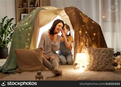 family, hygge and people concept - happy mother with little daughter whispering in kids tent at night at home. happy family whispering in kids tent at home