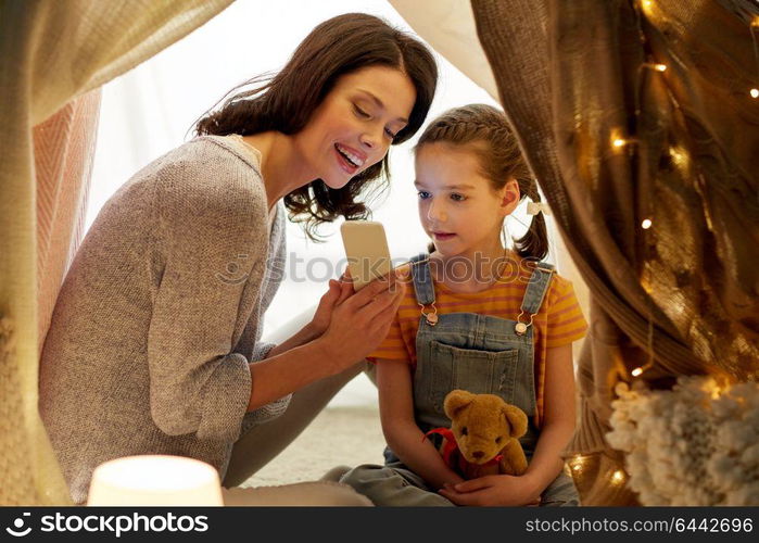 family, hygge and technology concept - happy mother and little daughter with smartphone in kids tent at night at home. happy family with smartphone in kids tent at home