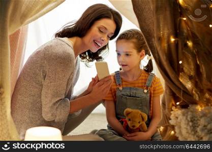 family, hygge and technology concept - happy mother and little daughter with smartphone in kids tent at night at home. happy family with smartphone in kids tent at home