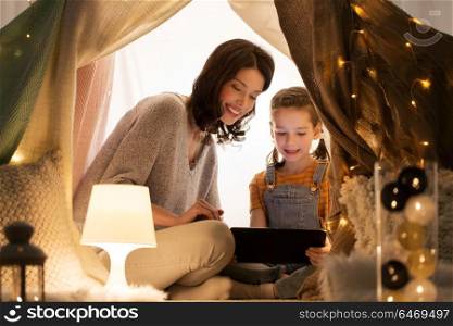 family, hygge and technology concept - happy mother and little daughter with tablet pc computer in kids tent at night at home. family with tablet pc in kids tent at home
