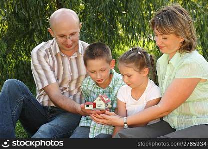 family in early fall park. father, mother, little boy and girl is sitting on the grass near osier and looking at wendy house.