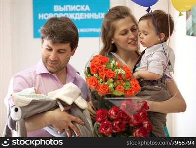 Family in the maternity hospital, father holds infant, mother with flowers holds elder son