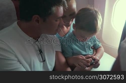 Family is on the plane, boy is sitting on mother&acute;s lap and playing a game in smartphone.