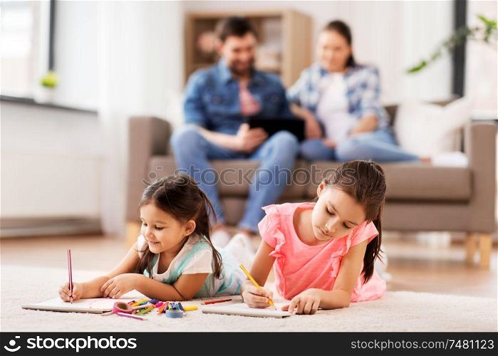 family, leisure and childhood concept - happy sisters lying on floor and drawing in sketchbooks with crayons at home. happy sisters drawing in sketchbooks at home