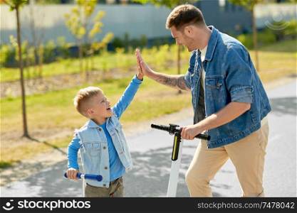 family, leisure and fatherhood concept - happy father spending time with little son riding scooters and making high five in city. father and son with scooters making high five