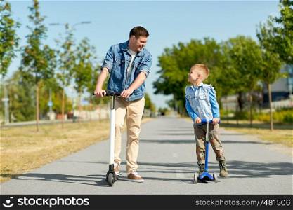 family, leisure and fatherhood concept - happy father spending time with little son riding scooters in city. father and little son riding scooters in city