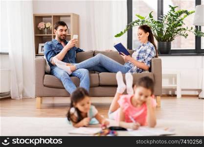family, leisure and people concept - happy daughters with crayons drawing in sketchbooks, mother and father with tablet pc computer sitting on sofa at home. happy family spending free time at home
