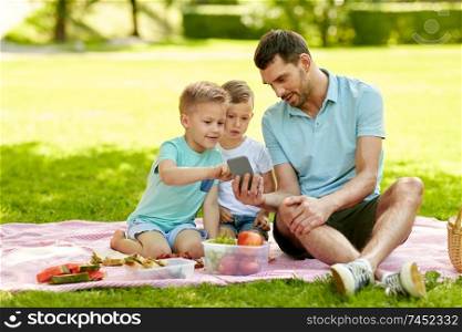 family, leisure and people concept - happy father and two little sons with smartphone having picnic at summer park. family with smartphone having picnic at park