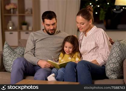 family, leisure and people concept - happy father, mother and little daughter reading book at home at night. happy family reading book at home at night