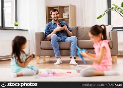 family, leisure and people concept - happy father with smartphone sitting on sofa and looking at his daughters doing crafts at home. father with smartphonew and daughters at home