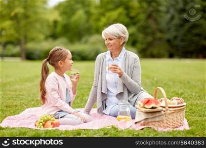 family, leisure and people concept - happy grandmother and granddaughter having picnic and drinking fruit water at summer park. grandmother and granddaughter at picnic in park