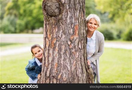 family, leisure and people concept - happy grandmother and granddaughter peeking out tree at summer park. grandmother and granddaughter behind tree at park