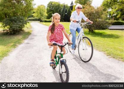 family, leisure and people concept - happy grandmother and granddaughter riding bicycles at summer park. grandmother and granddaughter cycling at park