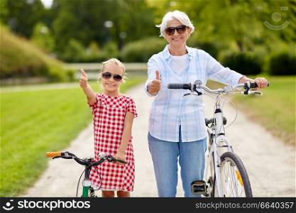 family, leisure and people concept - happy grandmother and granddaughter with bicycles showing thumbs up at summer park. grandmother and granddaughter with bicycles