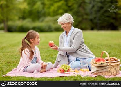 family, leisure and people concept - happy grandmother giving apple to granddaughter on picnic at summer park. grandmother and granddaughter at picnic in park