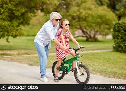 family, leisure and people concept - happy grandmother teaching granddaughter to ride bicycle at summer park. grandmother and granddaughter with bicycles