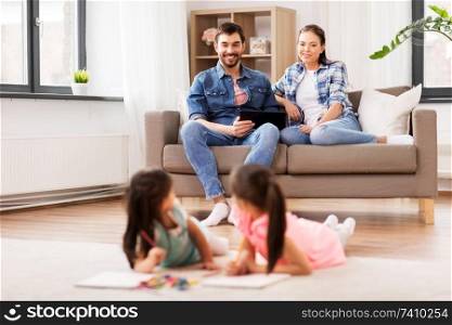 family, leisure and people concept - happy mother and father with tablet pc computer sitting on sofa and looking at daughters drawing at home. happy family spending free time at home