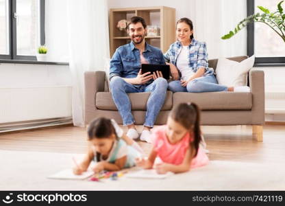 family, leisure and people concept - happy mother and father with tablet pc computer sitting on sofa and looking at daughters drawing at home. happy family spending free time at home