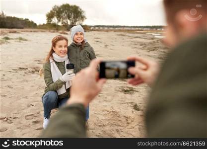 family, leisure and people concept - happy mother, father and little daughter photographing by smartphone on autumn beach. family photographing by smartphone on autumn beach