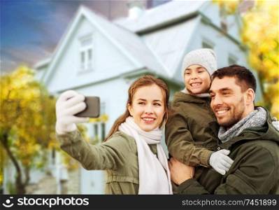 family, leisure and people concept - happy mother, father and little daughter taking selfie by smartphone over house in autumn background. family taking selfie over house in autumn