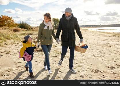family, leisure and people concept - happy mother, father and little son walking along autumn beach with picnic basket and blanket. happy family going to picnic on beach in autumn