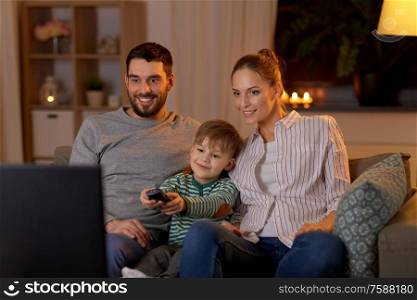 family, leisure and people concept - happy smiling father, mother and little son with remote control watching tv at home at night. happy family watching tv at home at night