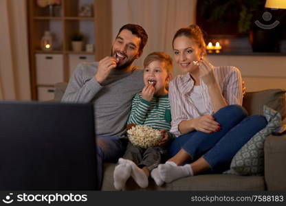 family, leisure and people concept - happy smiling father, mother and little son eating popcorn and watching tv at home in evening. happy family with popcorn watching tv at home