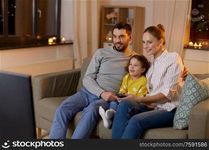 family, leisure and people concept - happy smiling father, mother and little daughter watching tv at home at night. happy family watching tv at home at night