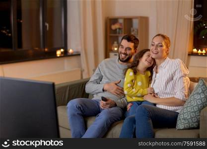 family, leisure and people concept - happy smiling father, mother and little daughter watching something funny on tv at home at night. happy family watching tv at home at night