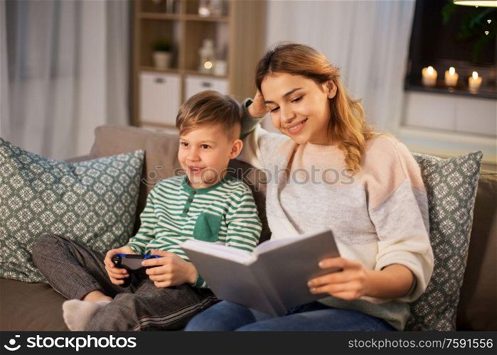family, leisure and people concept - happy smiling mother reading book and her little son with gamepad playing video game in evening. happy mother with little son spending time at home