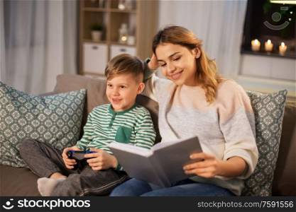 family, leisure and people concept - happy smiling mother reading book and her little son with gamepad playing video game in evening. happy mother with little son spending time at home