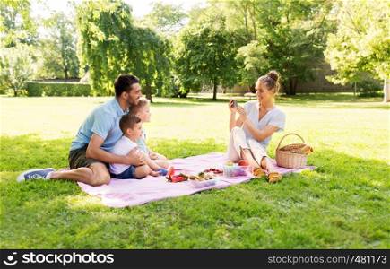 family, leisure and people concept - mother taking picture of happy father with two little sons by smartphone on picnic at summer park. mother taking picture of family on picnic at park