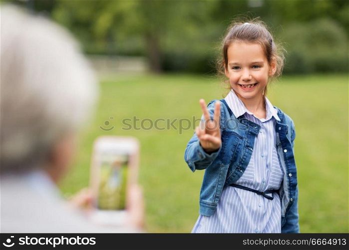 family, leisure and technology concept - happy little girl being photographed and showing peace hand sign at summer park. girl being photographed and showing peace sign