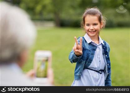family, leisure and technology concept - happy little girl being photographed and showing peace hand sign at summer park. girl being photographed and showing peace sign