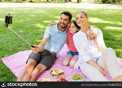 family, leisure and technology concept - happy mother, father and daughter having picnic and taking picture by smartphone on selfie stick at summer park. family having picnic and taking selfie at park