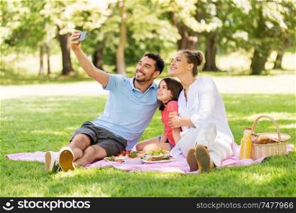 family, leisure and technology concept - happy mother, father and daughter having picnic and taking selfie by smartphone at summer park. family having picnic and taking selfie at park