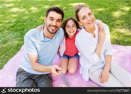 family, leisure and technology concept - happy mother, father and daughter having picnic and taking picture by selfie stick at summer park. family having picnic and taking selfie at park