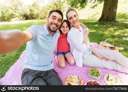 family, leisure and technology concept - happy mother, father and daughter having picnic and taking selfie at summer park. family having picnic and taking selfie at park