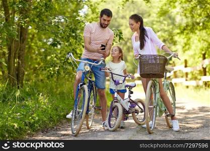 family, leisure and technology concept - happy mother, father and little daughter with smartphone and bicycles in summer park. family with smartphone and bicycles in summer park