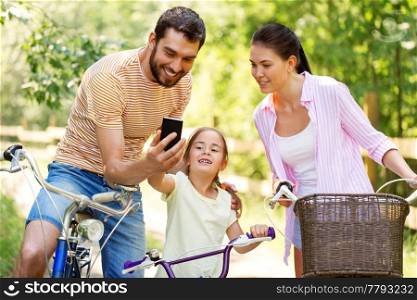 family, leisure and technology concept - happy mother, father and little daughter with smartphone and bicycles in summer park. family with smartphone and bicycles in summer park