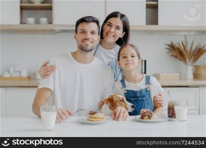Family, leisure, pastime, eating concept. Father, mother and daughter, jack russell terrier dog pose all together at camera against kitchen interior, get pleasure from eating pancakes drink fresh milk