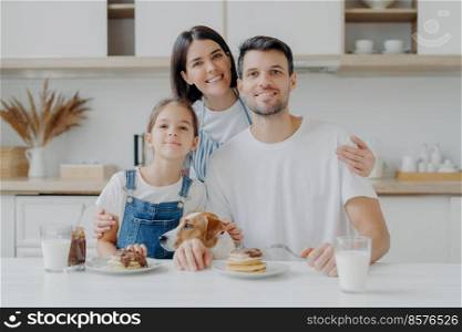 Family, leisure, pastime, eating concept. Father, mother and daughter, jack russell terrier dog pose all together at camera against kitchen interior, get pleasure from eating pancakes drink fresh milk