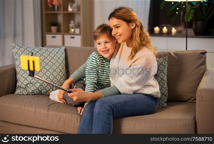family, love and people concept - happy smiling mother with little son taking picture by smartphone on selfie stick at home in evening. mother and son taking selfie by smartphone at home