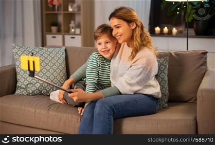 family, love and people concept - happy smiling mother with little son taking picture by smartphone on selfie stick at home in evening. mother and son taking selfie by smartphone at home
